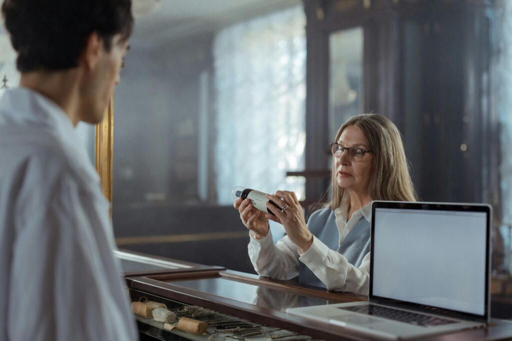 Senior woman helps customer with medication query at a pharmacy counter indoors.